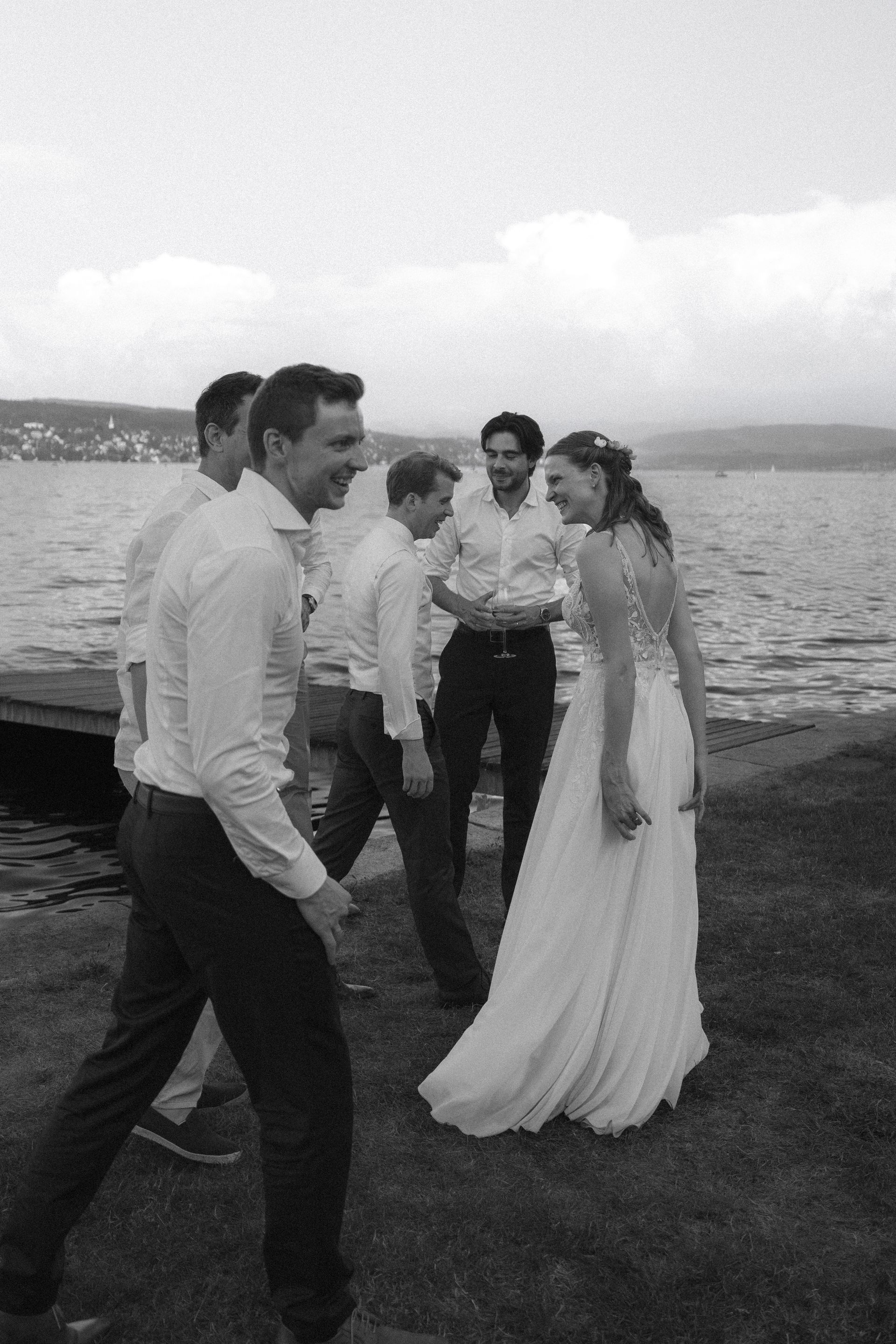 A bride and groom walking a lakeside promenade with their wedding guests, in black and white