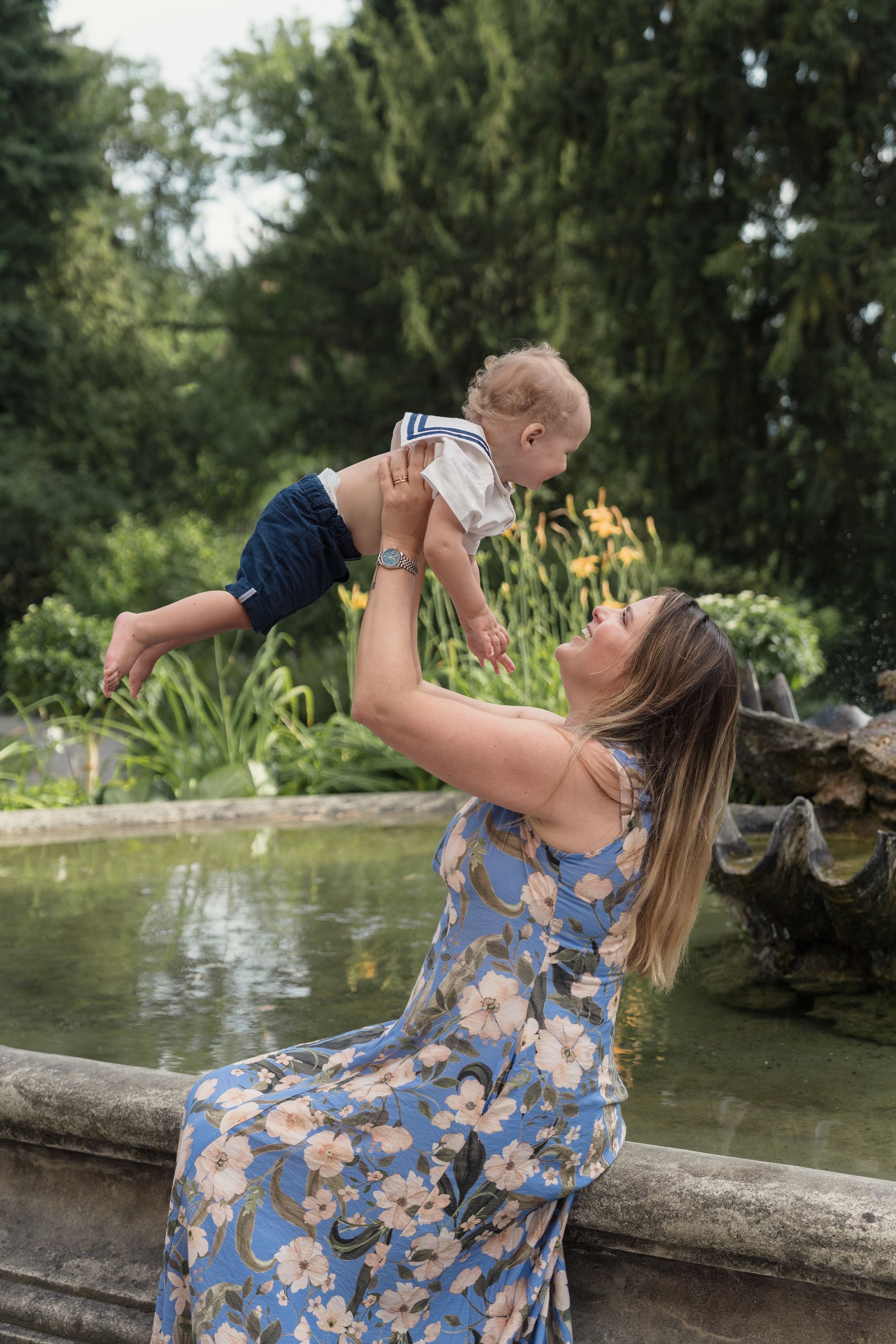A mother in a floral dress lifting her baby above a garden pond at Villa Patumbah, Zürich