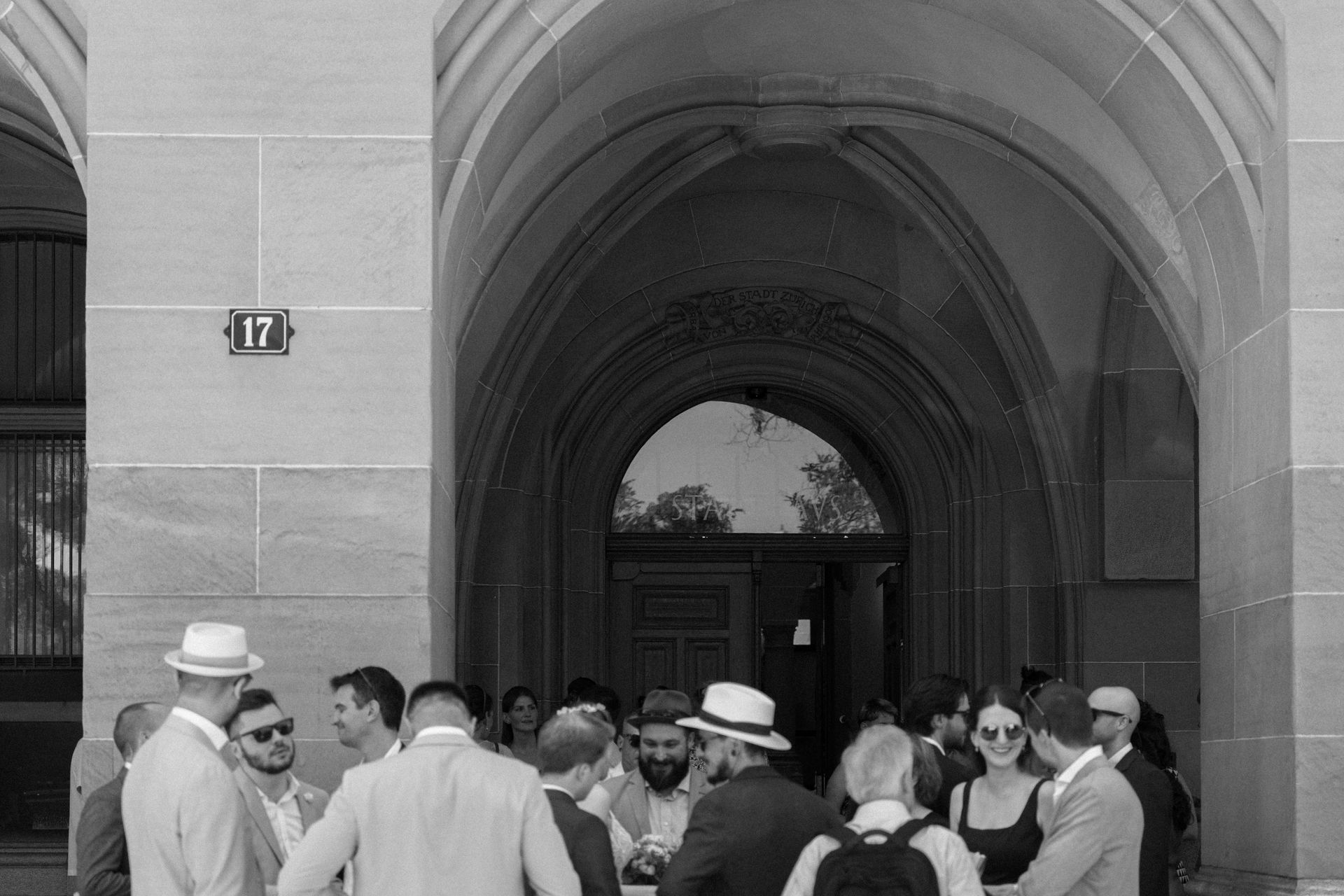 Wedding guests gathered under a stone archway outside the Stadthaus register office in Zürich, in black and white