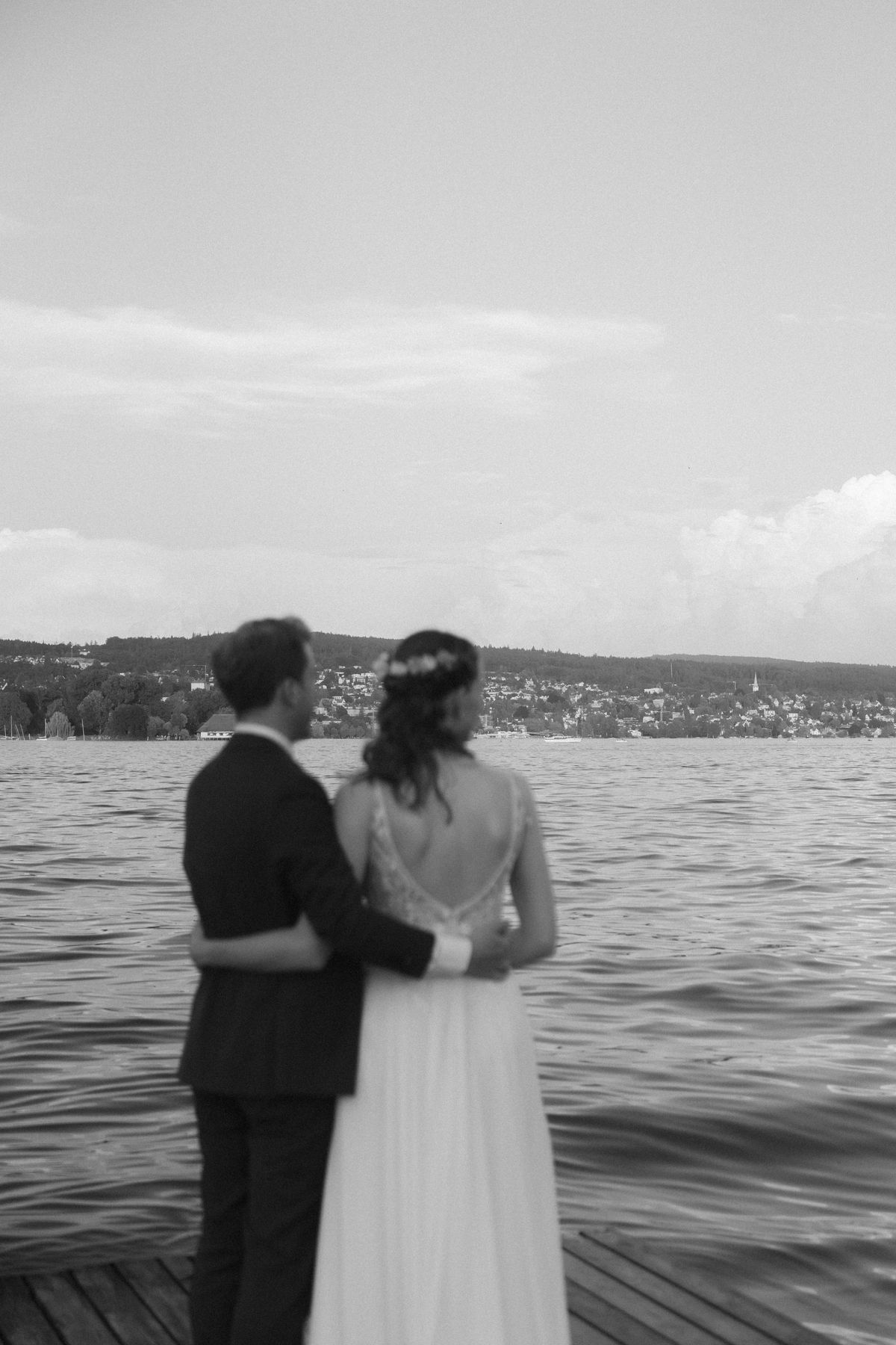 A newlywed couple holding each other, looking out over Lake Zürich